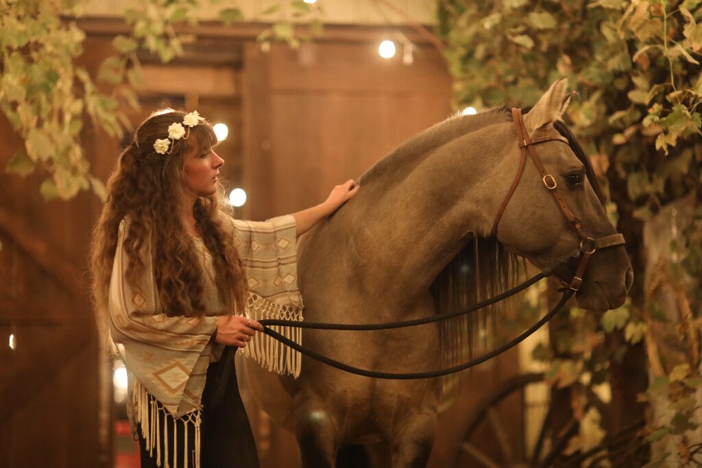 Girl wearing flower crown and shawl petting a grulla Kiger Mustang under grape arbor with wooden barn doors