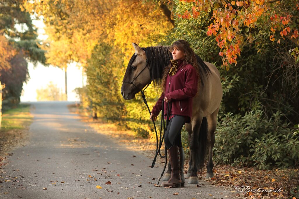 Gold and orange tree lined lane with girl in a purple jacket holding a grulla Kiger Mustang in a rope halter
