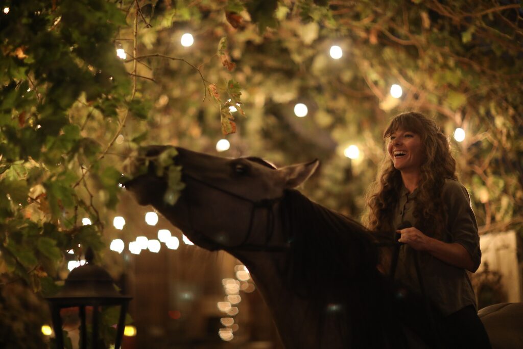 Girl laughs on horseback as her grulla Kiger mustang tries to nibble leaves from the lit grape arbor arching over them